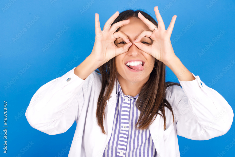 Young european doctor woman on blue background doing ok gesture like binoculars sticking tongue out, eyes looking through fingers. Crazy expression. Young european doctor woman on blue background doing ok gesture like binoculars sticking tongue out, eyes looking through fingers. Crazy expression.