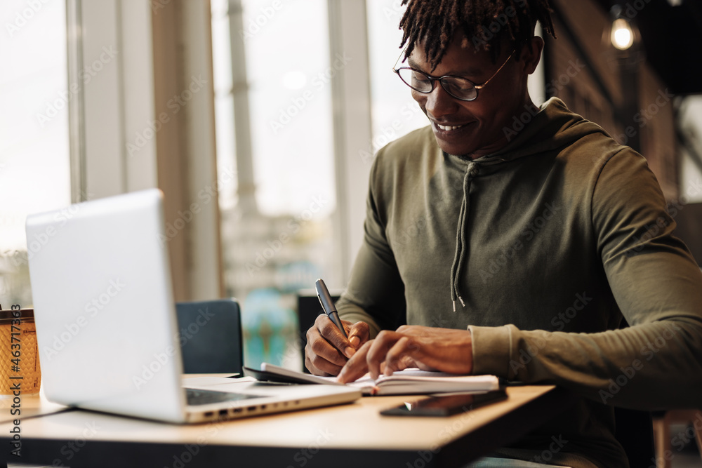 African American man with laptop and notepad at the table. a man writes with a pen in a notebook. distance learning African American man with laptop and notepad at the table. a man writes with a pen in a notebook. distance learning