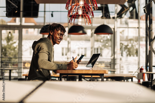 an African American man with a laptop and a phone in his hands, sitting at a table in the office, writes in a notebook photo
