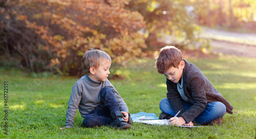 The kid is watching as the older brother next to him doing his homework sitting on the grass