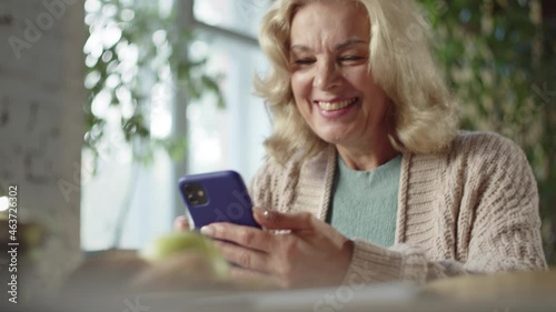 A woman aged sitting at a table and watching a video on the phone. She smiles sweetly and laughs