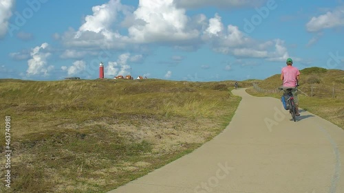 Slow motion of a bicyle path in the dunes. people riding bicycles through the beuautifull landscape. In the distance we see a lighthouse in the dunes. The sky is blue, clouds passing by. Serie of 7 photo