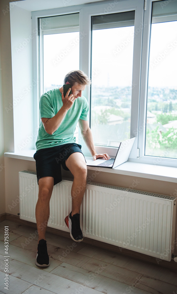 Side view of a man sitting on the windowsill talking on the phone with his co-worker while looking for plane tickets for a vacation at sea on his laptop Side view of a man sitting on the windowsill talking on the phone with his co-worker while looking for plane tickets for a vacation at sea on his laptop