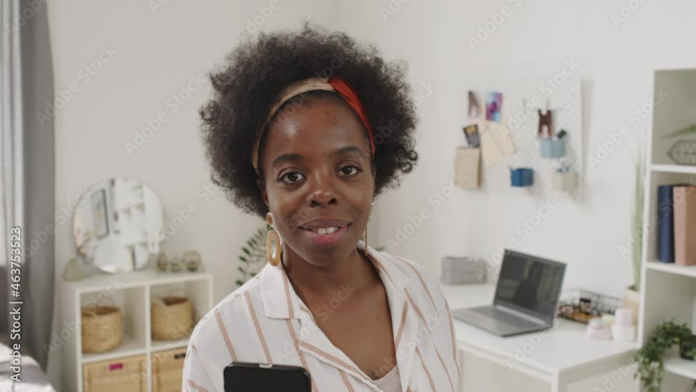 Medium close-up portrait with slowmo of smiling young African-American woman posing for camera standing at bright modern apartment holding smartphone Medium close-up portrait with slowmo of smiling young African-American woman posing for camera standing at bright modern apartment holding smartphone