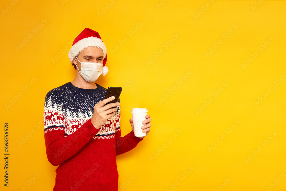 man in medical mask, Christmas sweater, red Santa Claus hat holding paper cup with hot drink and phone. writes reads message with congratulations, chooses gifts for new year. concept app, online, sale man in medical mask, Christmas sweater, red Santa Claus hat holding paper cup with hot drink and phone. writes reads message with congratulations, chooses gifts for new year. concept app, online, sale