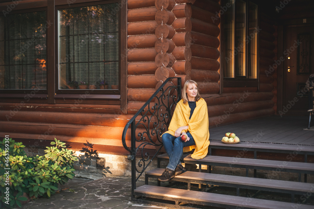 A beautiful blonde woman sits on the steps of the porch of a large wooden house. The woman is wrapped in a blanket, smiling and holding an apple in her hand. A place for text.