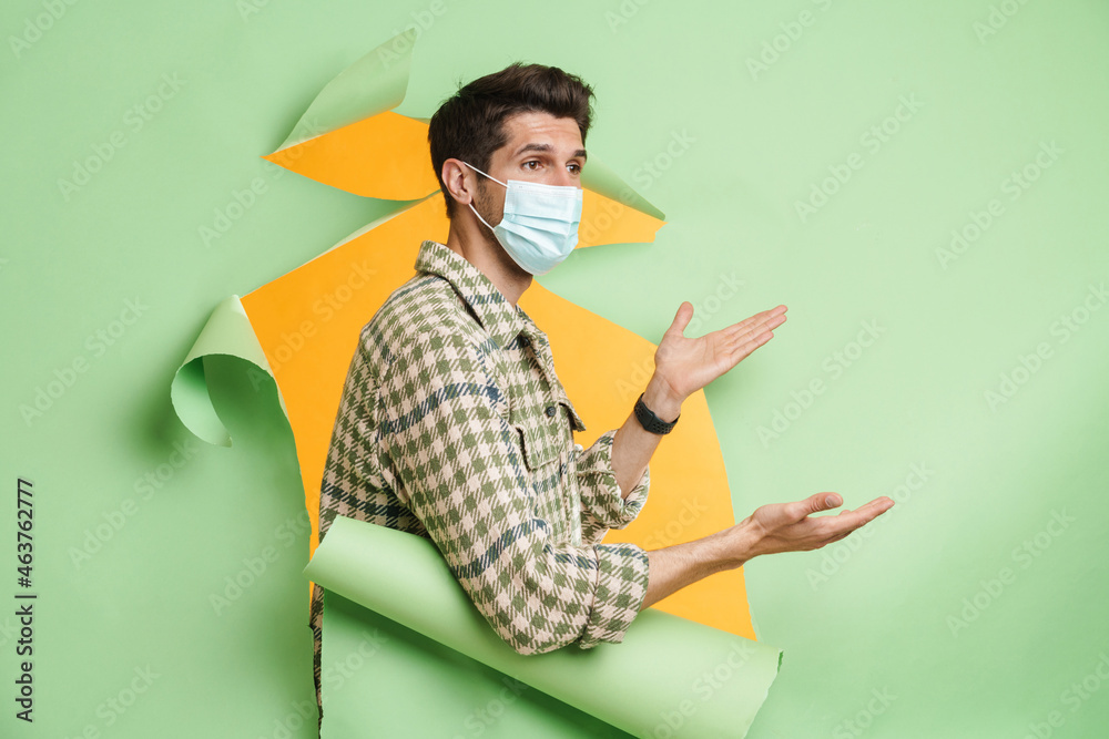 Man wearing face mask gesturing while posing into hole in green background Man wearing face mask gesturing while posing into hole in green background