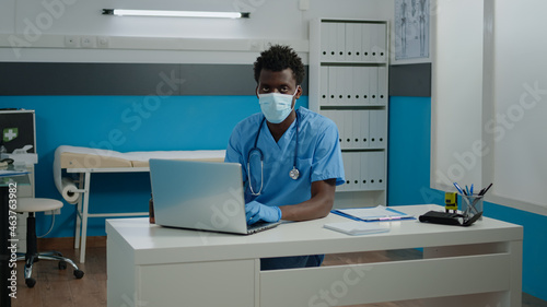 Portrait of man with nurse profession wearing uniform at face mask looking at camera in healthcare office. Young adult sitting at desk while typing on laptop keyboard during pandemic