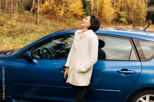 Happy smiling fashion business woman in white female coat with cell phone laughing outside her car in autumn park. Win concept © MarijaBazarova