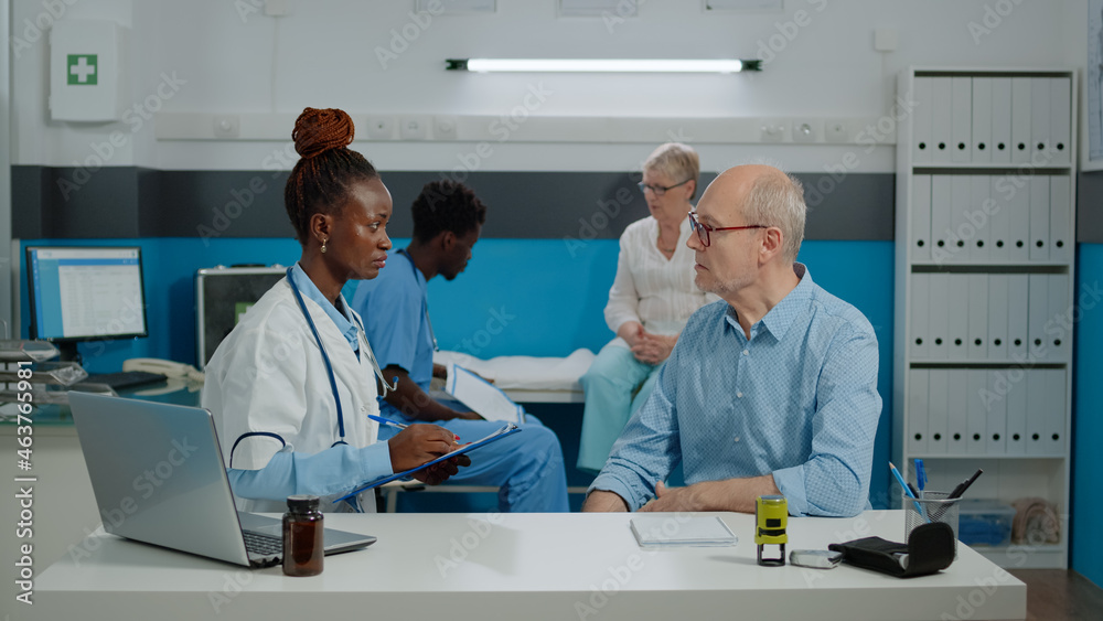 Senior woman receiving medical advice from man nurse while sitting on bed in cabinet at facility. Young doctor talking to elder patient at desk while writing checkup information