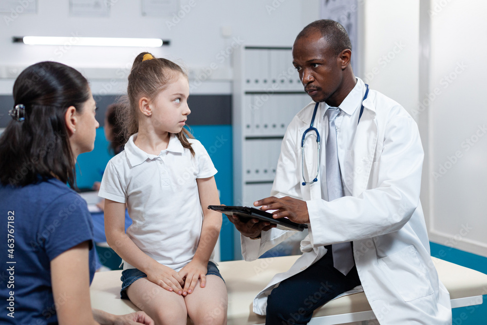 Obraz premium African american pediatrician doctor typing disease expertise of young child on tablet computer while discussing medical treatment during clinical appointment in hospital office. Health care service