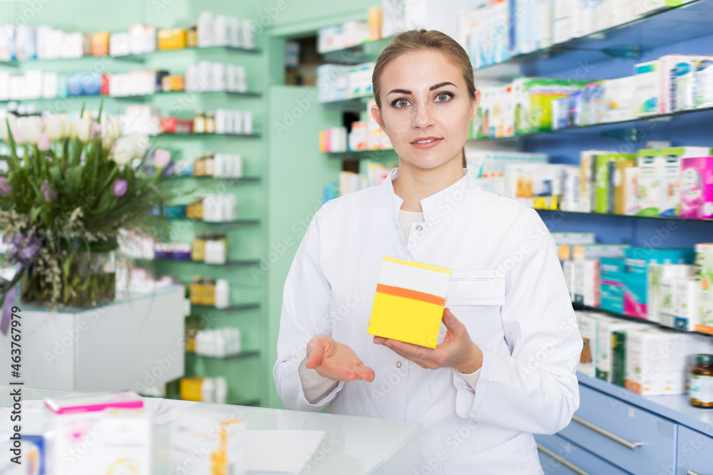Ordinary woman pharmacist is standing welcoming near cashbox in pharmacy
