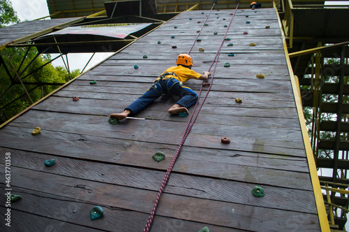 A nine-year-old boy equipped with a rock-climbing gear kit is climbing a wall outdoor with confidence (low angle) photo