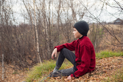 A boy in the autumn forest sits and looks thoughtfully into the distance.