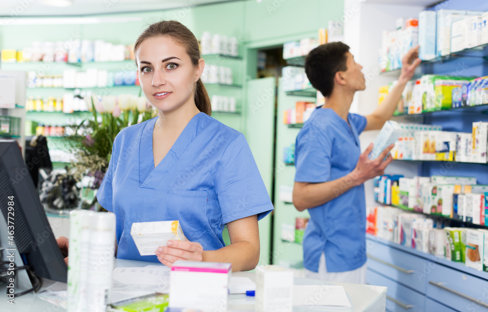 positive female and male specialist are attentively stocktaking medicines with notebook near shelves in pharmacy.