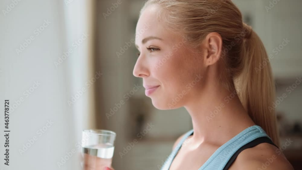 Side view of smiling blond woman drinking water at home near the window