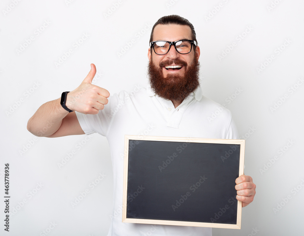 Bearded man wearing glasses is showing thumb up and holding a black chalk board. Bearded man wearing glasses is showing thumb up and holding a black chalk board.