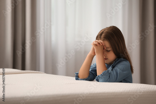Cute little girl with hands clasped together saying bedtime prayer in bedroom. Space for text