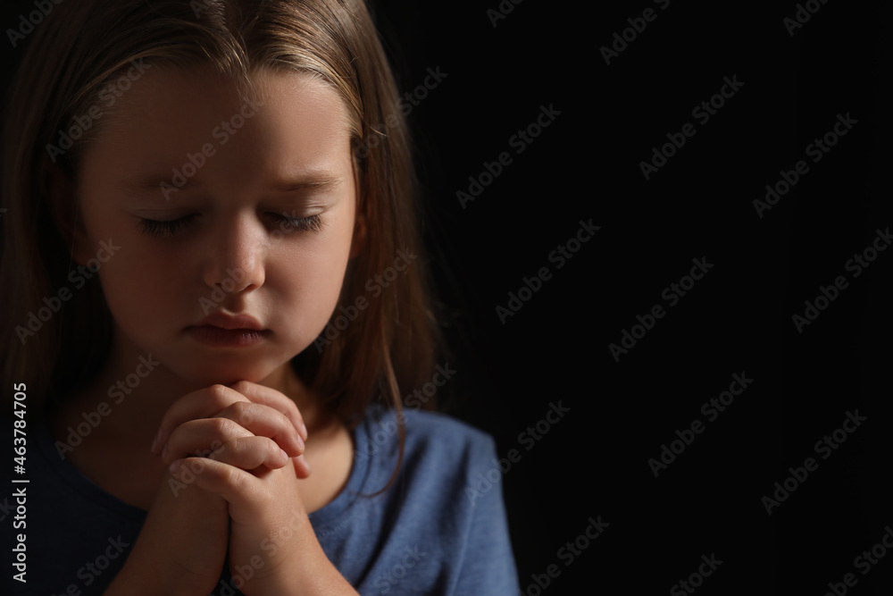 Cute little girl with hands clasped together praying on black background. Space for text