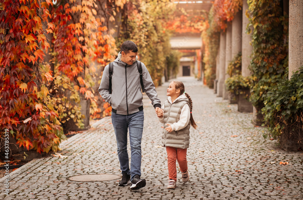 Fototapeta premium Young dad and little daughter walk together by the hands in the autumn park. Family time, togehterness, parenting and happy childhood concept. Weekend with emotions.