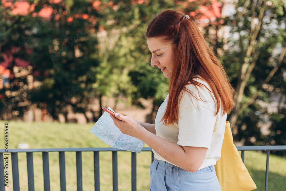 Naklejka premium Attractive young female tourist is exploring new city. Happy redhead Woman with map and smartphone in hands lost in new place, relaxation or traveling concept