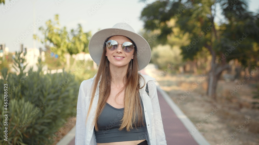 Happy young woman in summer clothes, hat and sunglasses walking with backpack outdoors. Palm trees around. Concept of people, vacation and tourism.
