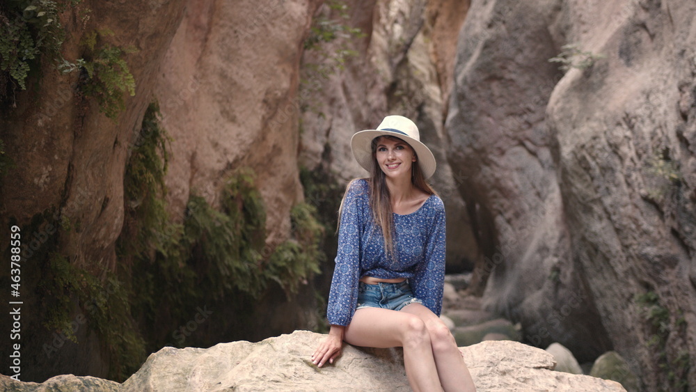 Stunning young woman sitting on a large rock among the rocks and smiling at the camera. Pretty Caucasian woman wearing summer hat and clothes. Enjoy summer vacation Stunning young woman sitting on a large rock among the rocks and smiling at the camera. Pretty Caucasian woman wearing summer hat and clothes. Enjoy summer vacation
