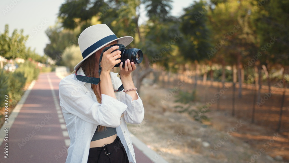 Young woman photographer wanderer. Happy young woman using modern digital camera for taking photos while walking. Female tourist in summer hat and clothes saving memories from trip