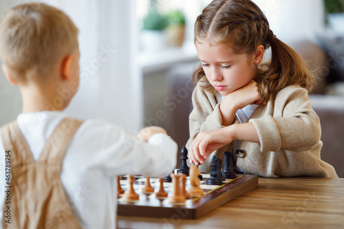 Focused children playing chess game at home while sitting in living room at table with chessboard photo