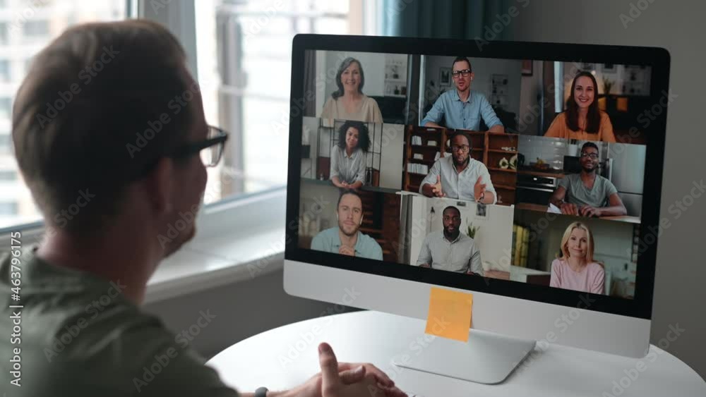 Attentive young hipster man sitting at table with PC, working remotely from home, on team meeting online with coworkers, looking at screen with people profiles and waving hand