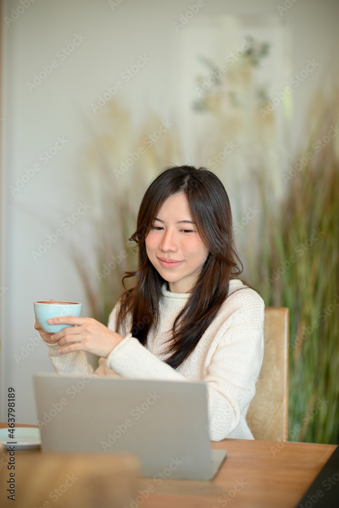 Beautiful millennial asian woman drinking hot coffee while working on laptop computer at cafe. Beautiful millennial asian woman drinking hot coffee while working on laptop computer at cafe.