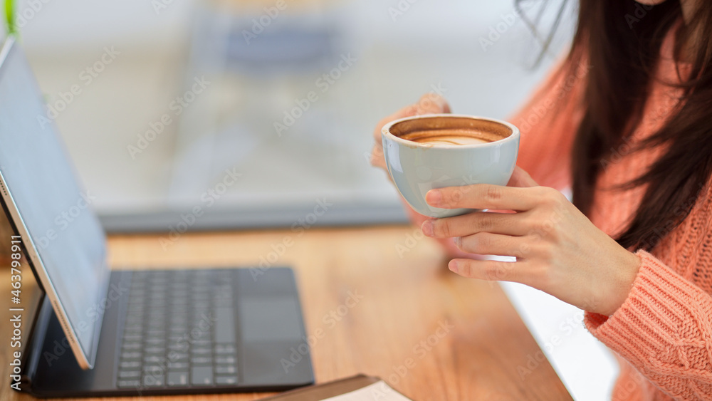 Young female in pink sweater drinking, holding hot coffee cup while working on tablet Young female in pink sweater drinking, holding hot coffee cup while working on tablet