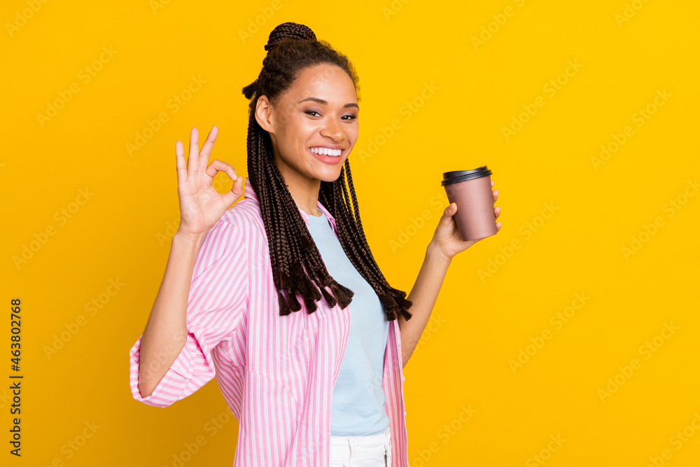 Photo of young happy positive dark skin woman make okay sign hold coffee cup wear striped shirt isolated on yellow color background Photo of young happy positive dark skin woman make okay sign hold coffee cup wear striped shirt isolated on yellow color background
