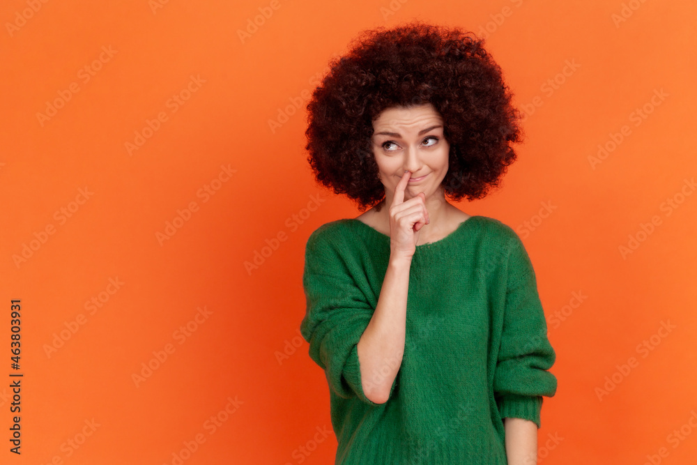 Funny woman with Afro hairstyle wearing green casual style sweater picking her nose, demonstrating bad manners, childish behavior. Indoor studio shot isolated on orange background. Funny woman with Afro hairstyle wearing green casual style sweater picking her nose, demonstrating bad manners, childish behavior. Indoor studio shot isolated on orange background.