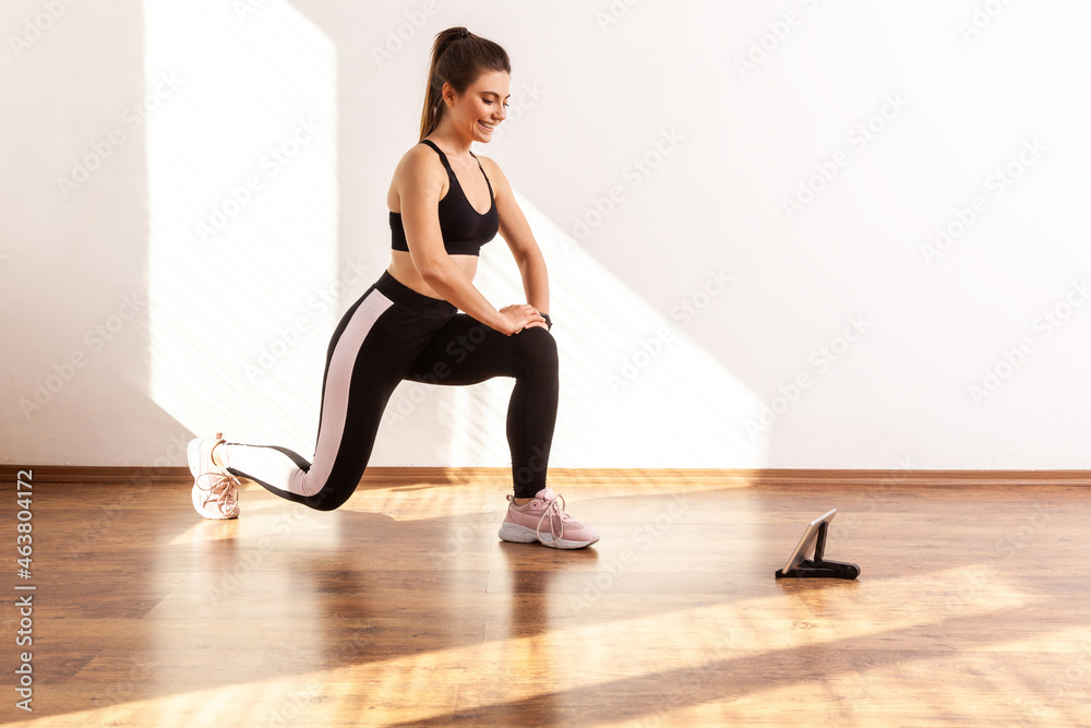 Girl doing lunge or split squats, repeating after fitness blogger, watching training video on tablet, wearing black sports top and tights. Full length studio shot illuminated by sunlight from window.