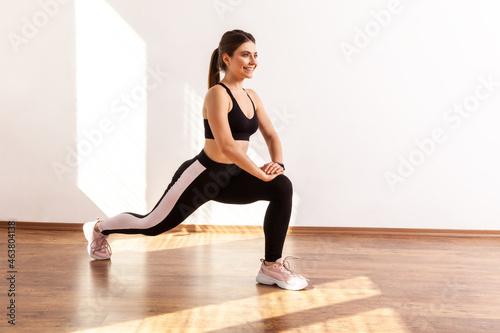 Slim athletic beautiful female stretching legs before workout, lower body exercise, wearing black sports top and tights. Full length studio shot illuminated by sunlight from window.