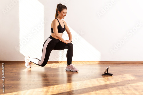 Girl doing lunge or split squats, repeating after fitness blogger, watching training video on tablet, wearing black sports top and tights. Full length studio shot illuminated by sunlight from window.