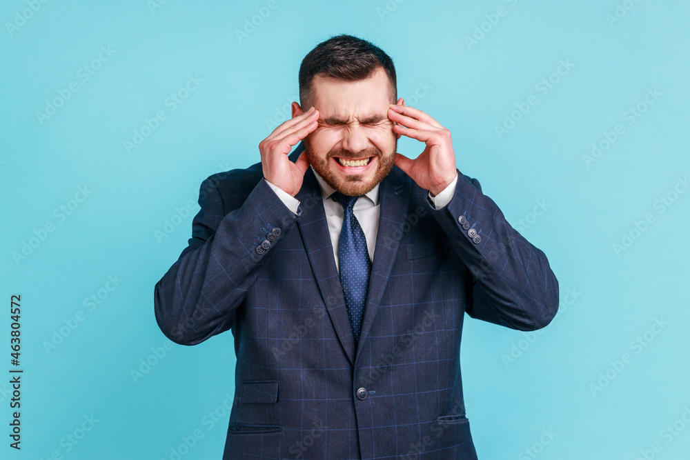 Unhappy frustrated bearded man wearing official style suit massaging temples feeling headache, suffering migraine or high blood pressure. Indoor studio shot isolated on blue background. Unhappy frustrated bearded man wearing official style suit massaging temples feeling headache, suffering migraine or high blood pressure. Indoor studio shot isolated on blue background.