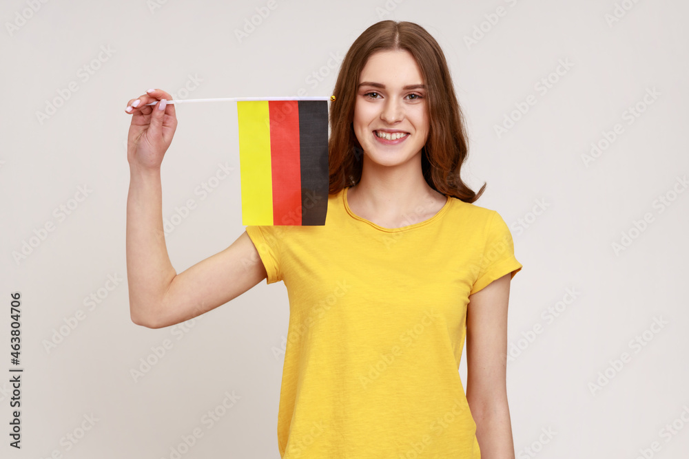 Portrait of pleasant looking teenager girl in yellow T-shirt holding Germany flag, celebrating Day of Germany - 3th October, expressing positive. Indoor studio shot isolated on gray background. Portrait of pleasant looking teenager girl in yellow T-shirt holding Germany flag, celebrating Day of Germany - 3th October, expressing positive. Indoor studio shot isolated on gray background.