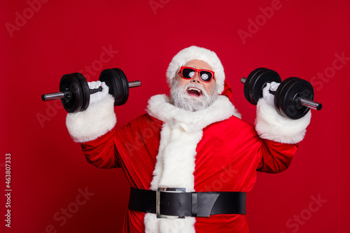 Portrait of attractive bearded cheery funny Santa lifting dumbbell working out isolated over bright red color background