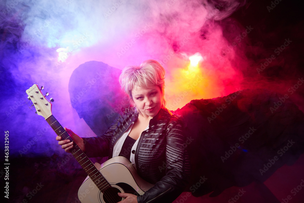 Self-assured adult blonde middle age woman with black angel wings and guitar like a rock musician. Model posing in studio on black background. Dangerous cupid on Valentine's day