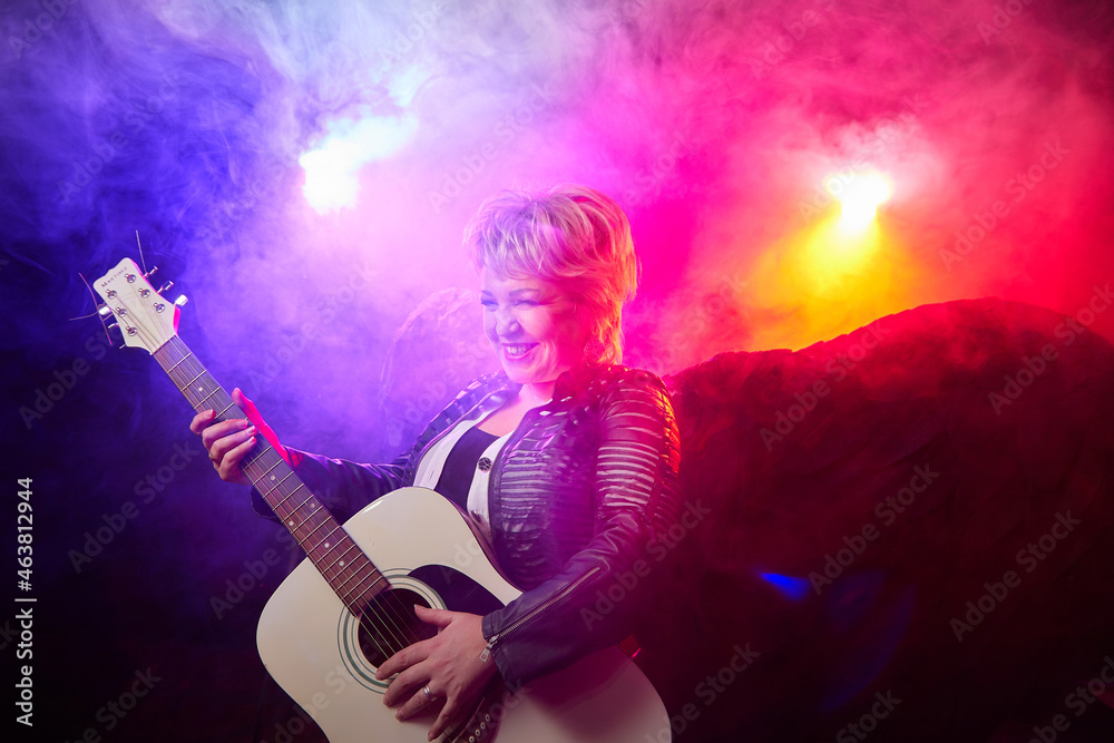 Obraz premium Self-assured adult blonde middle age woman with black angel wings and guitar like a rock musician. Model posing in studio on black background. Dangerous cupid on Valentine's day