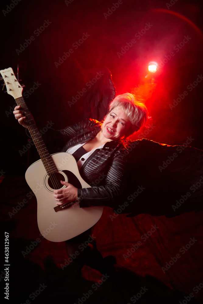 Self-assured adult blonde middle age woman with black angel wings and guitar like a rock musician. Model posing in studio on black background. Dangerous cupid on Valentine's day Self-assured adult blonde middle age woman with black angel wings and guitar like a rock musician. Model posing in studio on black background. Dangerous cupid on Valentine's day