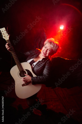 Self-assured adult blonde middle age woman with black angel wings and guitar like a rock musician. Model posing in studio on black background. Dangerous cupid on Valentine's day