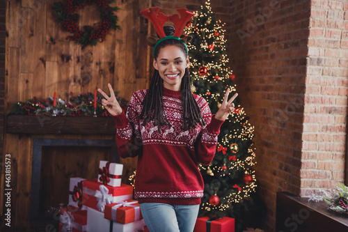 Photo of friendly positive lady have x-mas eve fun show v-sign wear horns jumper in decorated home indoors