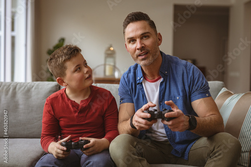 Caucasian father and son playing video games together sitting on the couch at home