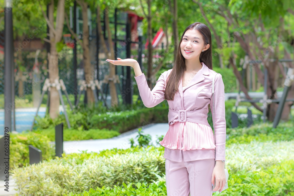 Beautiful young Asian professional business woman with long hair wearing pink shirt is smiling outdoor in garden front of the office while spreading hands to present something with trees background. Beautiful young Asian professional business woman with long hair wearing pink shirt is smiling outdoor in garden front of the office while spreading hands to present something with trees background.