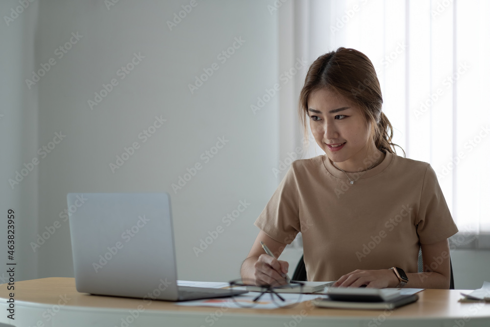 portrait of smiling woman making notes while taking part in webinar at tabletop with laptop in office portrait of smiling woman making notes while taking part in webinar at tabletop with laptop in office