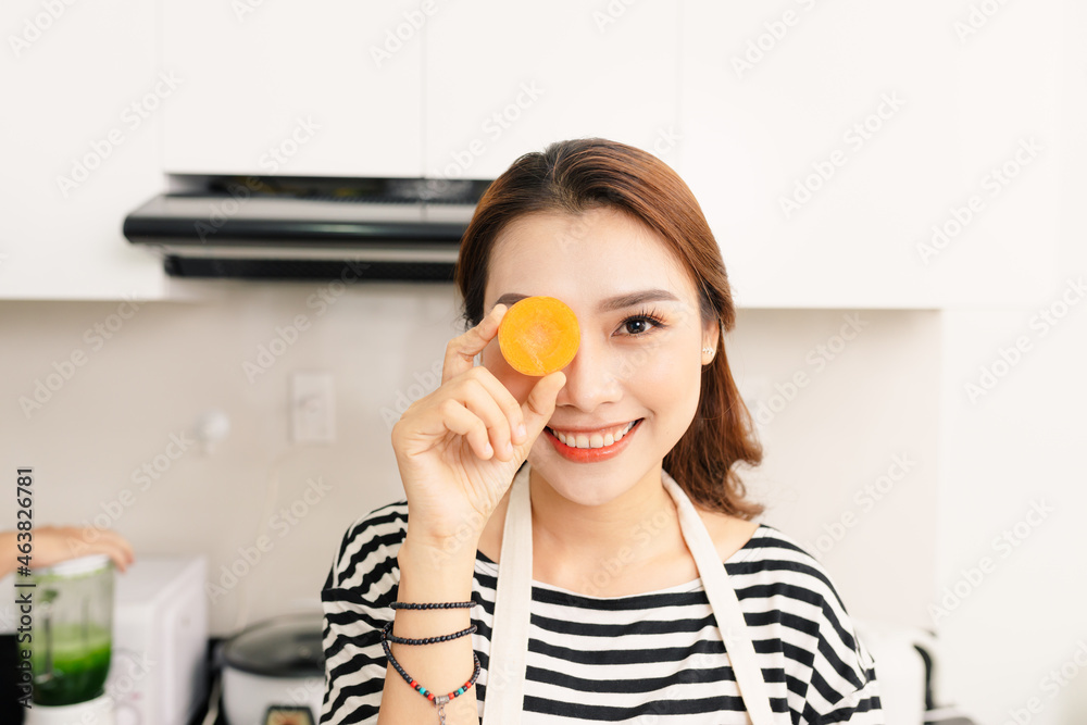 Young beautiful woman in orange apron sitting at table, having fun, carrot on eye Young beautiful woman in orange apron sitting at table, having fun, carrot on eye