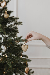 Women's hand hang decorations on beautiful Christmas tree with golden baubles and toys in front of white wall. Elegant aesthetic Christmas, New Year holidays celebration preparation background